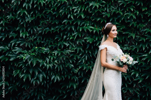 Portrait of a bride in a white dress with a wedding bouquet in the hands, against a background of a green wall of plants photo