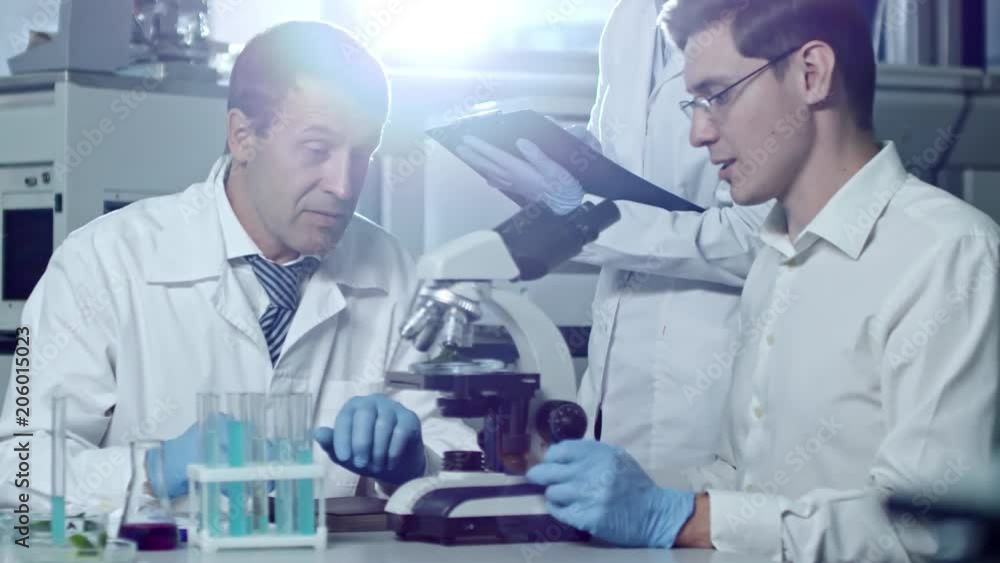 Medium shot of three chemists in lab coats studying samples with microscope when working in laboratory, woman writing down details Medium shot of three chemists in lab coats studying samples with microscope when working in laboratory, woman writing down details