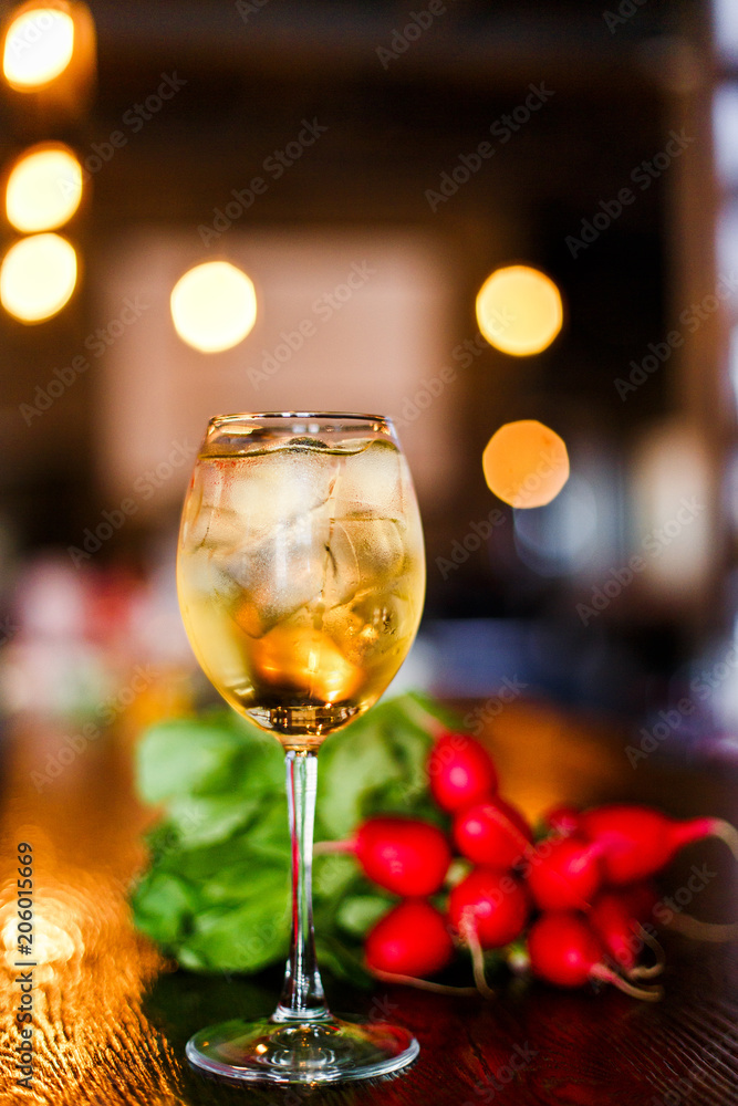 A photo of a light yellow cold summer aperitif alcoholic cocktail in a wine glass, full of ice cubes, garnished with radish. White wooden table. A photo of a light yellow cold summer aperitif alcoholic cocktail in a wine glass, full of ice cubes, garnished with radish. White wooden table.
