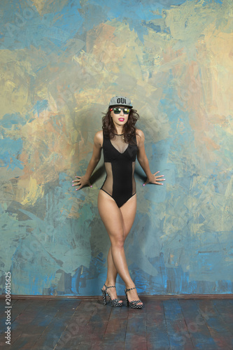 Vogue style Sexy slender young woman with a sporty body and an attractive breast with curly hair and red lips, with a cap and glasses in a black bodysuit in high heels with bare legs posing in a