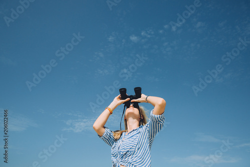 Beautiful natural blonde woman looking through binoculars against blue sky