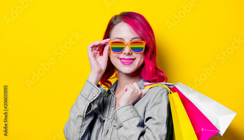 Young pink hair girl in rainbow sunglasses and blue shirt holding a colored shopping bags. Portrait on isolated yellow background