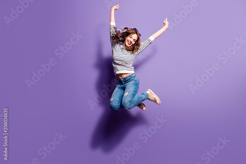 Fototapeta Portrait of cheerful positive girl jumping in the air with raised fists looking at camera isolated on violet background