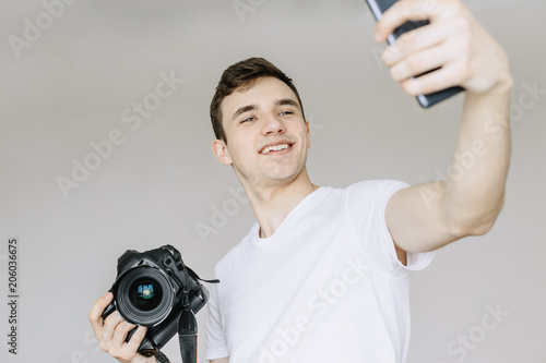 A young man holds a photo camera in his hand and makes a selfie by phone. Іsolated gray background photo