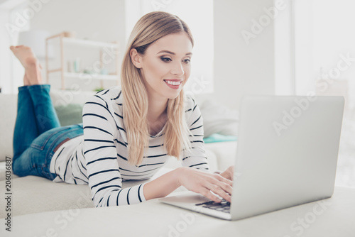 Portrait of pretty, charming, attractive, stylish, cheerful girl in jeans, striped outfit, having laptop, chatting with friends, using wi-fi internet, checking email, laying on sofa in livingroom