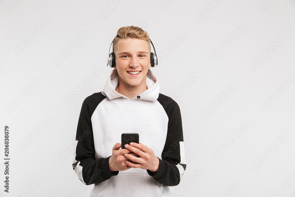 Portrait of teenage boy 16-18 years old wearing hoodie smiling and listening to music on smartphone via wireless headphones, isolated over white background Portrait of teenage boy 16-18 years old wearing hoodie smiling and listening to music on smartphone via wireless headphones, isolated over white background