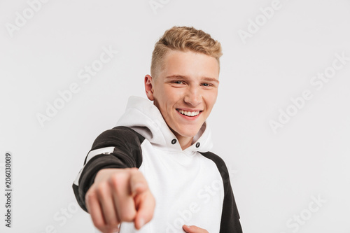 Portrait closeup of cheerful teenage boy 16-18 years old wearing hoodie pointing finger at camera with happy smile, isolated over white background