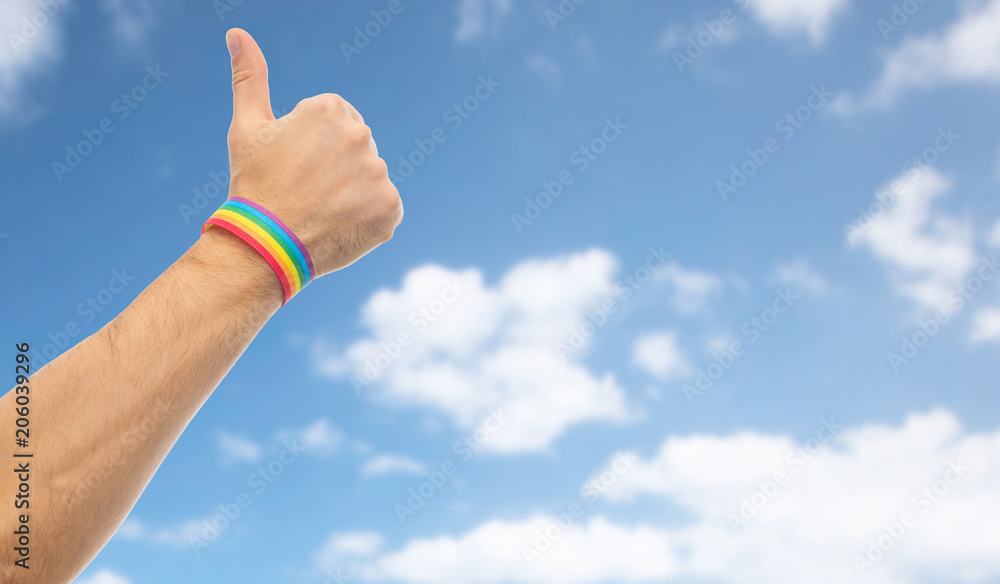 lgbt, same-sex relationships and homosexual concept - close up of male hand wearing gay pride awareness wristband showing thumbs up over blue sky and clouds background lgbt, same-sex relationships and homosexual concept - close up of male hand wearing gay pride awareness wristband showing thumbs up over blue sky and clouds background