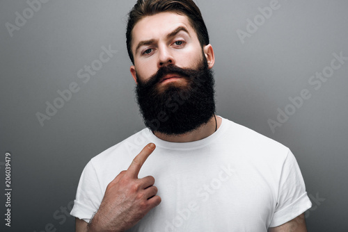 Clsoe up cropped portrait of brutal handsome Caucasian male indicate to his trendy beard, posing on gray studio background. Bearded barber European man model with confident expression on his face photo