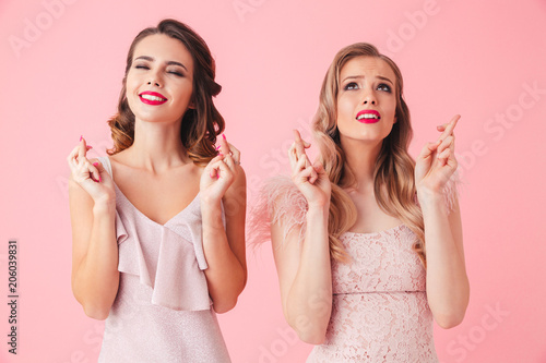 Two worried smiling women in dresses praying with crossed fingers