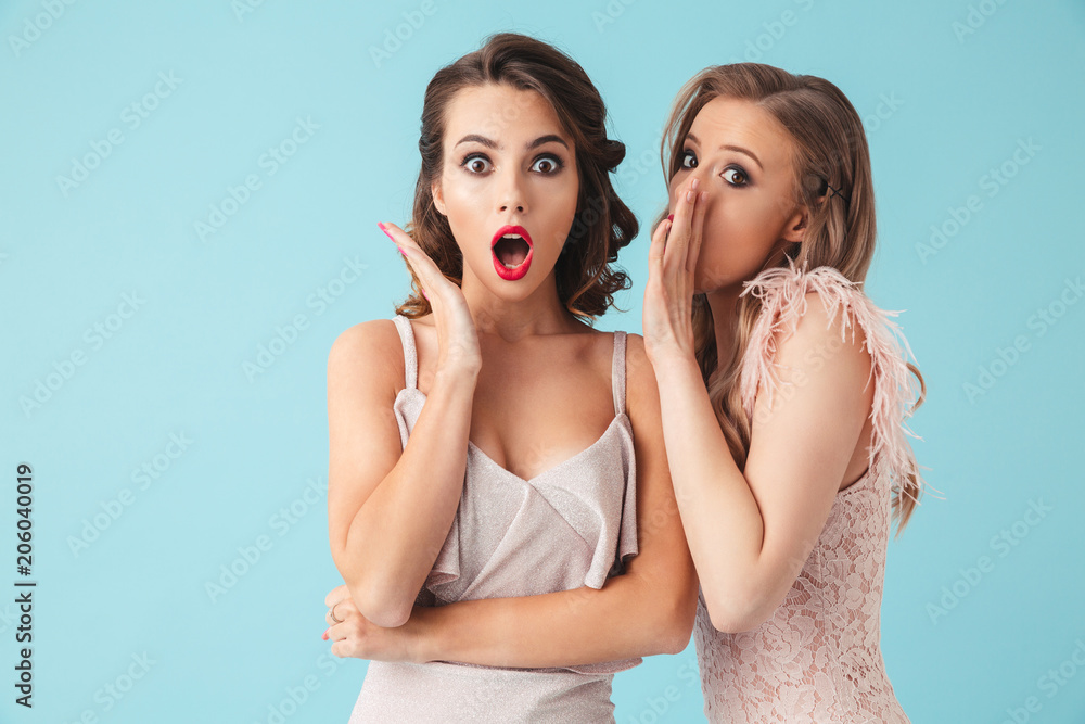 Photo of two gossip girls 20s wearing dresses and party makeup talking secrets in ear and expressing surprise, isolated over blue background Photo of two gossip girls 20s wearing dresses and party makeup talking secrets in ear and expressing surprise, isolated over blue background