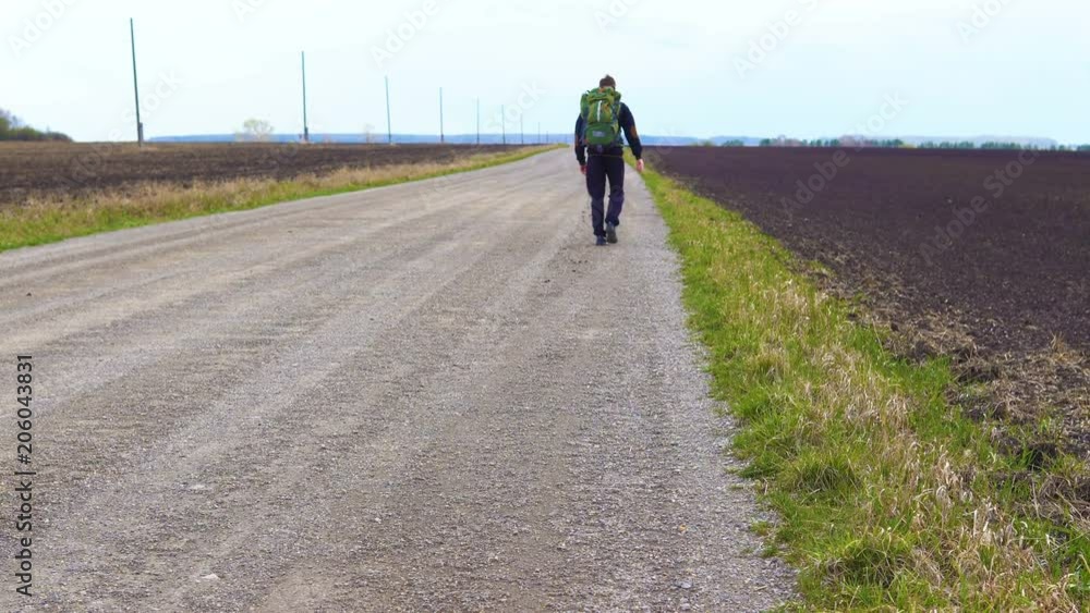 A young tourist walks along a dirt road along the fields with a backpack on his shoulders. Strong wind blows. A young tourist walks along a dirt road along the fields with a backpack on his shoulders. Strong wind blows.