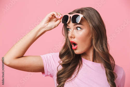 Photo closeup of shocked woman 20s looking aside at copyspace with open mouth from under fashionable stylish sunglasses, isolated over pink background photo