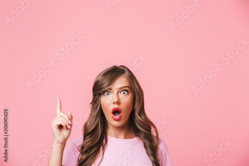 Closeup photo of excited woman with long curly brown hair pointing finger upward at copyspace with interest, isolated over pink background