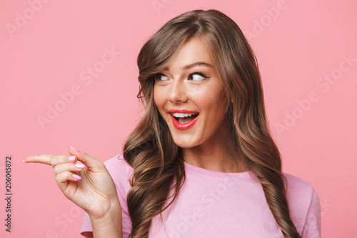 Closeup photo of adorable brunette woman in basic clothing pointing finger aside at copyspace with beautiful smile, isolated over pink background