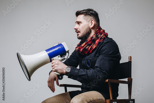 Young attractive movie director holding a megaphone. Film director talking on a loudspeaker while sitting on a chair isolated on grey background. Side view