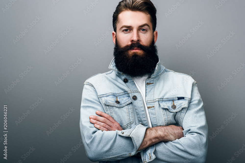 © iuricazac - Brutal handsome fashionable young European male with thick beard wearing trendy denim jacket, cross hands, staring at camera with serious and cheerful look. Barber man posing against gray studio wall.