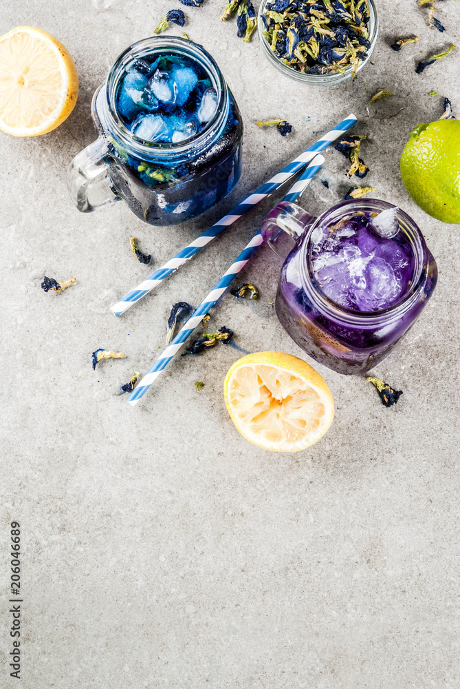 Healthy summer cold beverage, iced organic blue and violet butterfly pea flower tea with limes and lemons, grey concrete background copy space top view