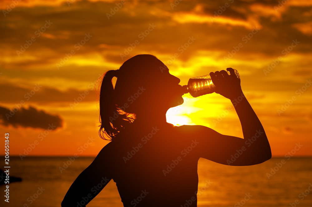 Silhouette of sportswoman - drinking water on warm summer evening during sunset