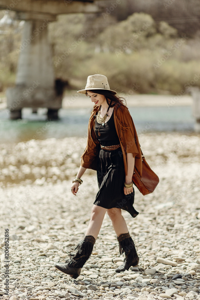 stylish hipster woman in hat, fringe poncho and boots walking on river beach. boho traveler girl in gypsy look, summer travel. atmospheric moment.  space for text.