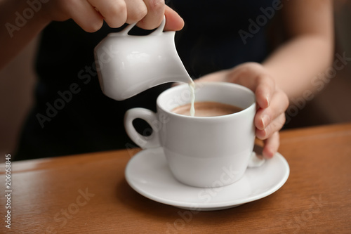 Woman pouring milk into cup of coffee on table