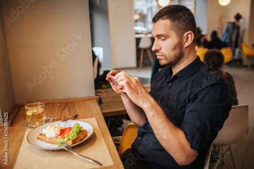Handsome man is sitting in cafe and taking a picture of meal he ordered. He is doing that on the phone. Guy looks very serious and thoughtful.
