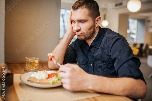 Bored and sad man is sitting at table and cafe. He is holding a piece of vegetable on fork. Man is looking at it and breathing out.