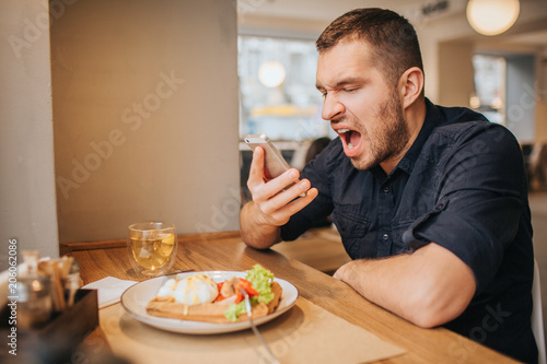 Mad man is sitting at table and screaming on the phone. He is angry and unhappy. Guy can't stop yelling. He doesn't eat food.