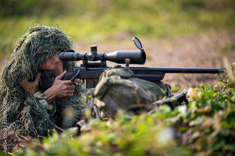 Fototapeta premium Sniper laying on the grass looking through scope at the target in deep forest.