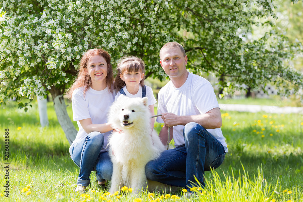 Happy family with a white dog in a summer park. The husband, wife and little daughter in white clothes and jeans, along with a white dog, rest in the garden. Happy family with a white dog in a summer park. The husband, wife and little daughter in white clothes and jeans, along with a white dog, rest in the garden.