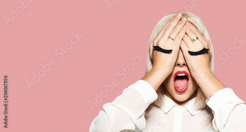 Young woman with gray and black eyelashes painted on hands. Blonde hair girl closed her eyes with hands and shout. Portrait of surprised model in white shirt. Concept of fun and tomfoolery.
