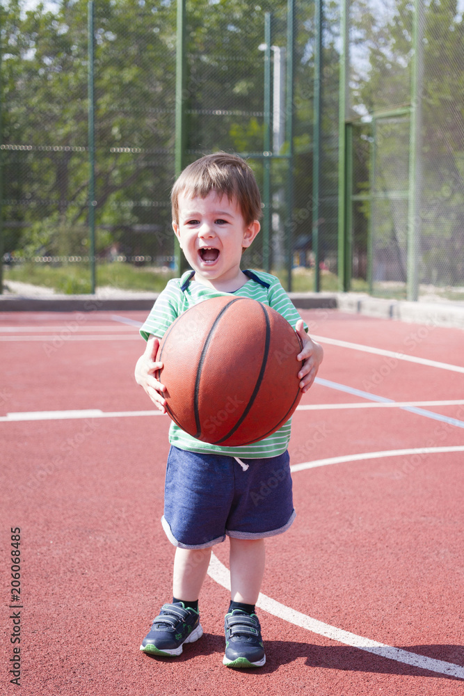 A boy half years playing with a ball on the sports field. Toddler is holding a big basketball balletable garden A boy half years playing with a ball on the sports field. Toddler is holding a big basketball balletable garden