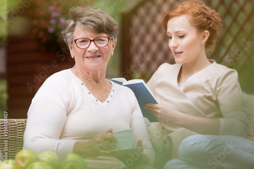 Принти на полотні Close-up of a happy senior woman and a her young professional nurse who is reading a book during leisure time on a patio of a day care facility