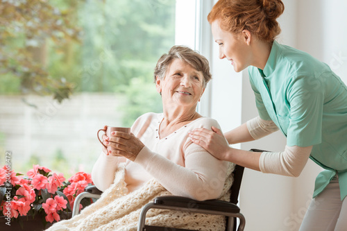 A professional caretaker in uniform helping a geriatric female patient on a wheelchair. Senior holding a cup and sitting by a large window in a rehabilitation center. photo