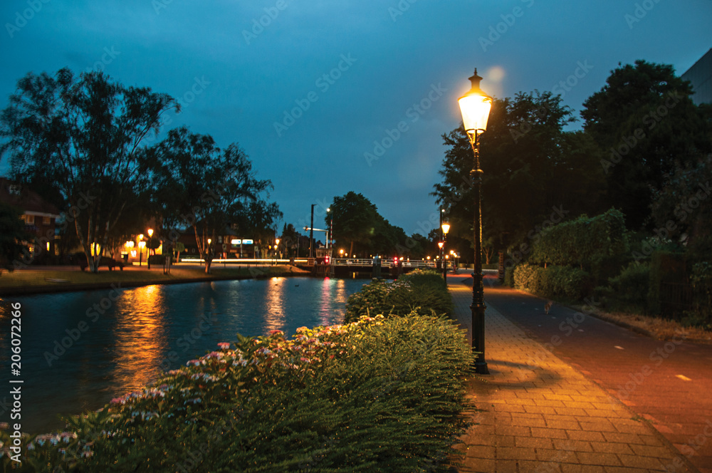 Night view of wide tree-lined canal, bridge and lamp post light in the foreground at dawn in Weesp. Quiet and pleasant village full of canals and green near Amsterdam. Northern Netherlands.