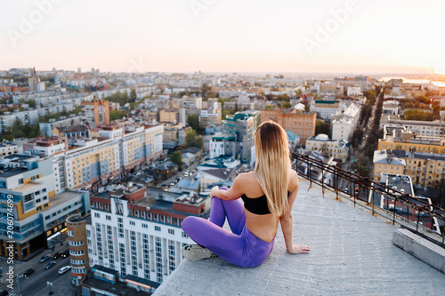 Young girl in sports uniform sits on the edge of the roof during sunset and looks to the horizon.