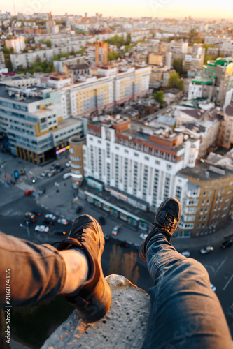 The guy is sitting on the edge of a tall building. He hung his legs with beautiful black sneakers over the city