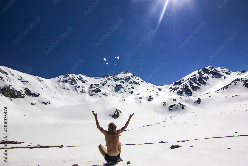 naked man sitting on snow field in mountains at daylight with hands up