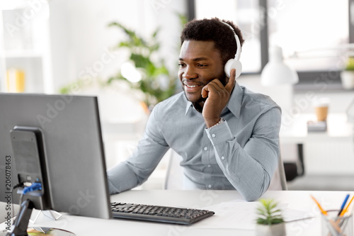 business, technology and people concept - happy african american businessman with headphones and computer computer listening to music at office