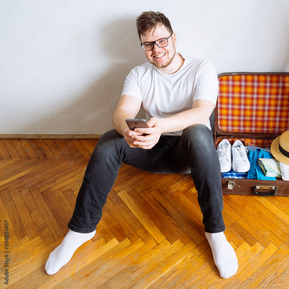 young man sit on floor checking list on mobile. valise packing for trip.travel concept. copy space. young man sit on floor checking list on mobile. valise packing for trip.travel concept. copy space.