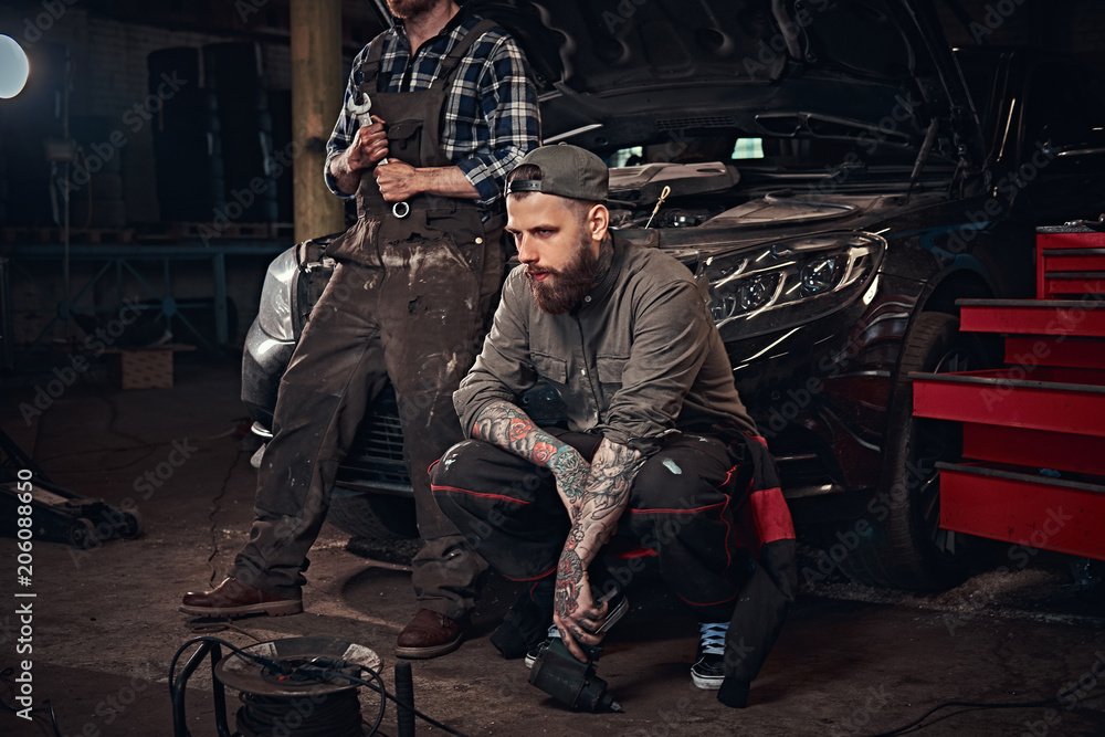Two mechanic sit on a broken car, rest after a hard day's work in a garage. Two mechanic sit on a broken car, rest after a hard day's work in a garage.
