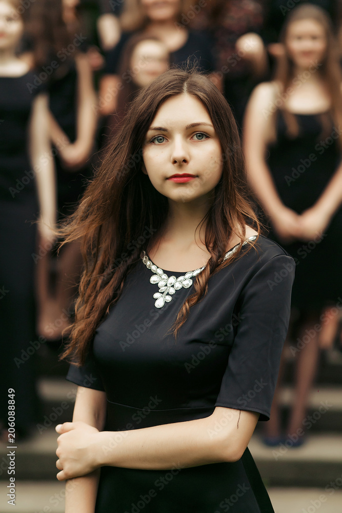 luxury gorgeous brunette woman in black dress posing, standing in front of group of ladys on background of stairs in city