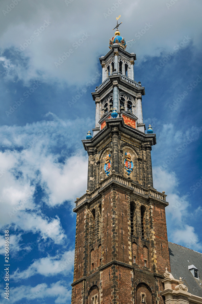 Close-up of pointed bell tower church made of bricks and golden clock under blue sky in Amsterdam. The city is famous for its huge cultural activity, graceful canals and bridges. Northern Netherlands.