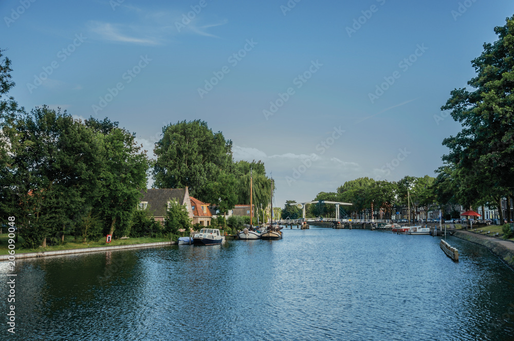 Calm river with grove and bridge, boats moored in the pier of brick houses and sunny blue sky in Weesp. Quiet and pleasant village full of canals and green near Amsterdam. Northern Netherlands.