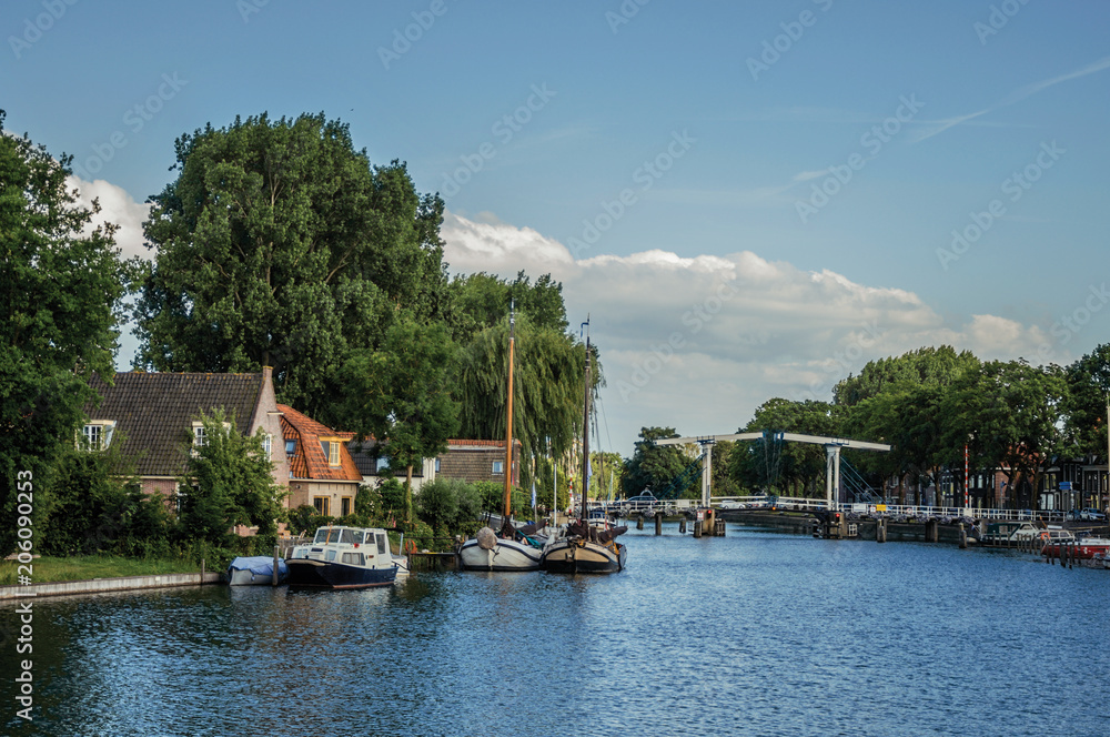 Calm river with grove and bridge, boats moored in the pier of brick houses and sunny blue sky in Weesp. Quiet and pleasant village full of canals and green near Amsterdam. Northern Netherlands.