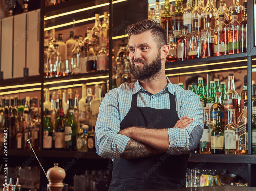 Stylish brutal bartender in a shirt and apron standing with crossed arms at bar counter background.
