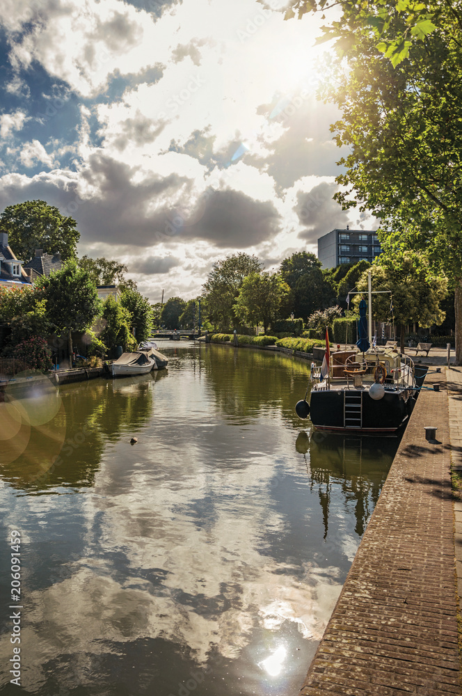 Fototapeta premium Wide canal with cloudy sky, houses and boat moored reflected in water on sunset at Weesp. Quiet and pleasant village full of canals and green near Amsterdam. Northern Netherlands. Retouched photo.
