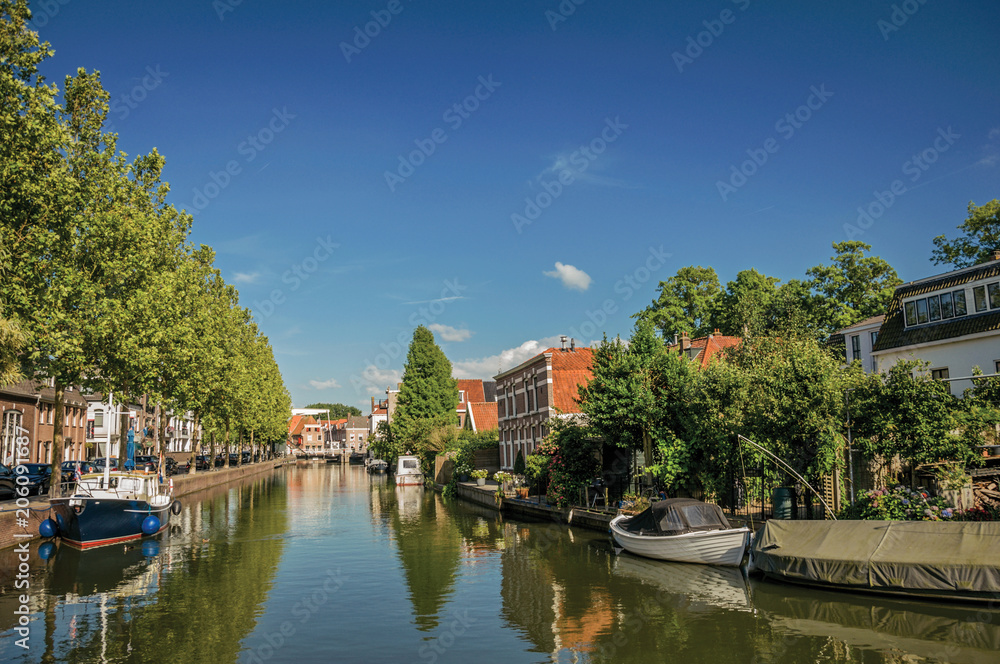 Wide canal with brick houses and boats moored on its bank reflected in water under blue sky of sunset at Weesp. Quiet and pleasant village full of canals and green near Amsterdam. Northern Netherlands