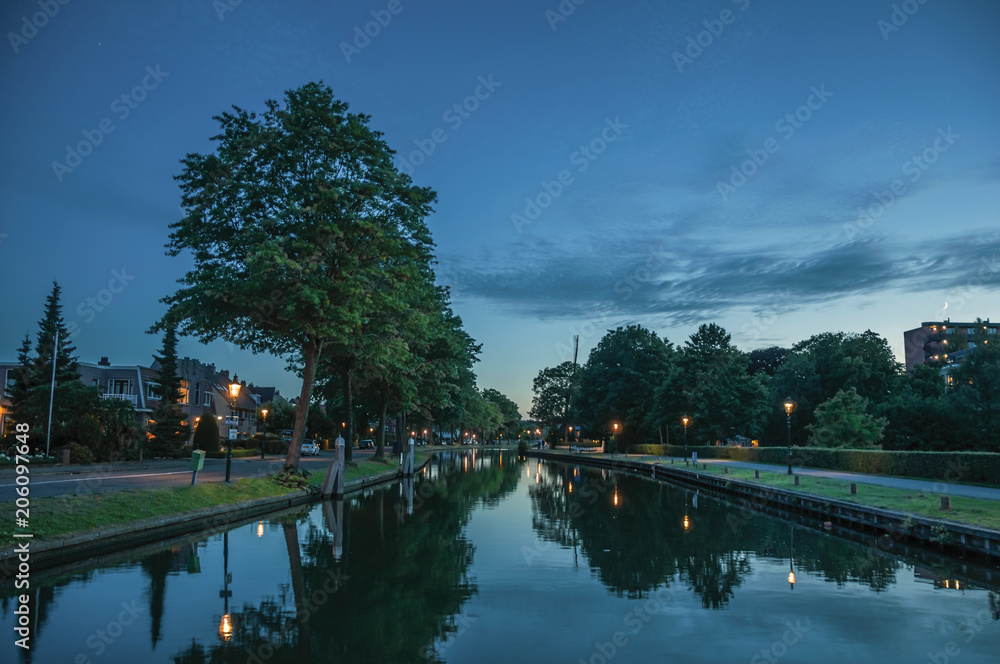 Night view of wide tree-lined canal, buildings and lamp post lighting at dawn in Weesp. Quiet and pleasant village full of canals and green near Amsterdam. Northern Netherlands. Night view of wide tree-lined canal, buildings and lamp post lighting at dawn in Weesp. Quiet and pleasant village full of canals and green near Amsterdam. Northern Netherlands.
