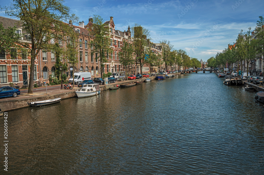 Tree-lined canal with old brick buildings, bridge, moored boats and sunny blue sky in Amsterdam. The city is famous for its huge cultural activity, graceful canals and bridges. Northern Netherlands.
